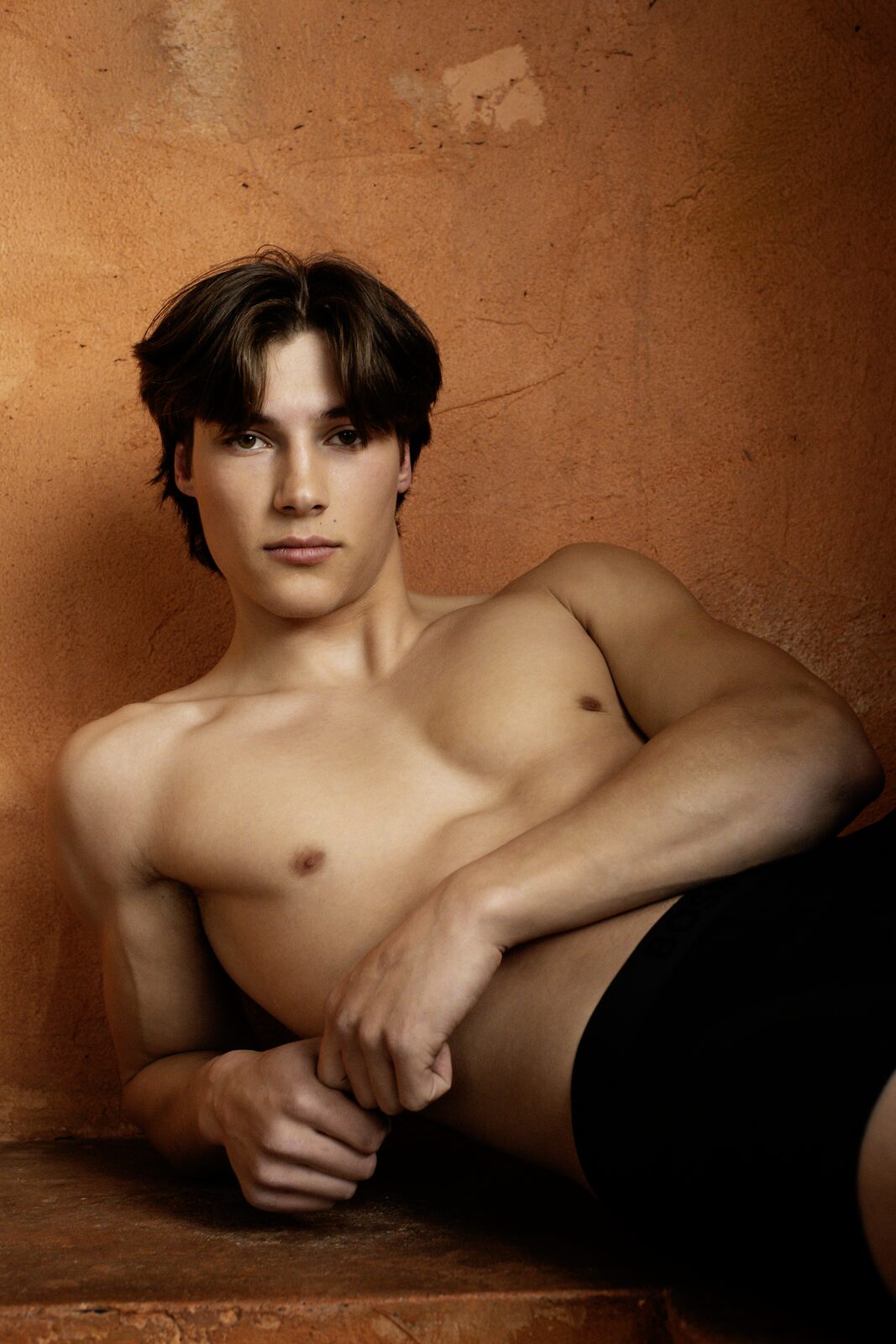 Male model reclining against terracotta wall, warm editorial portrait by Tony Liam