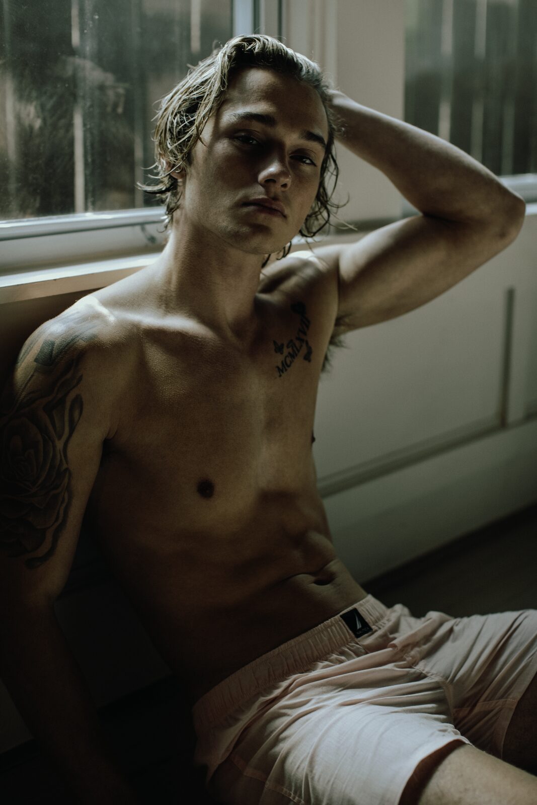 Moody portrait of tattooed male model with wet hair by window, by Tony Liam